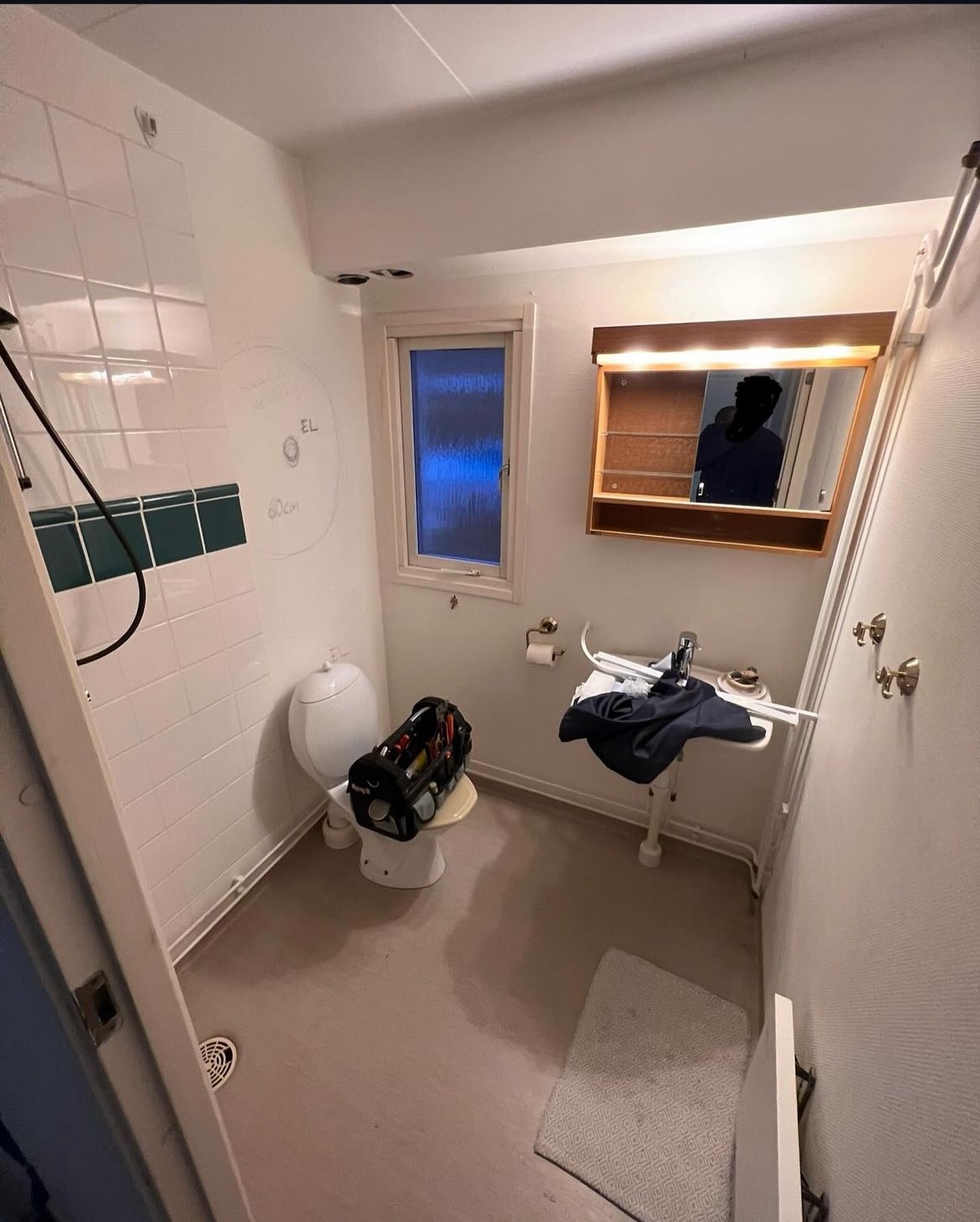 Łazienka w trakcie prac: narzędzia w torbie na sedesie, notatki na ścianie z glazurą, lustro z oświetleniem nad umywalką, małe okno z niebieską szybą, jasna podłoga i dywanik.