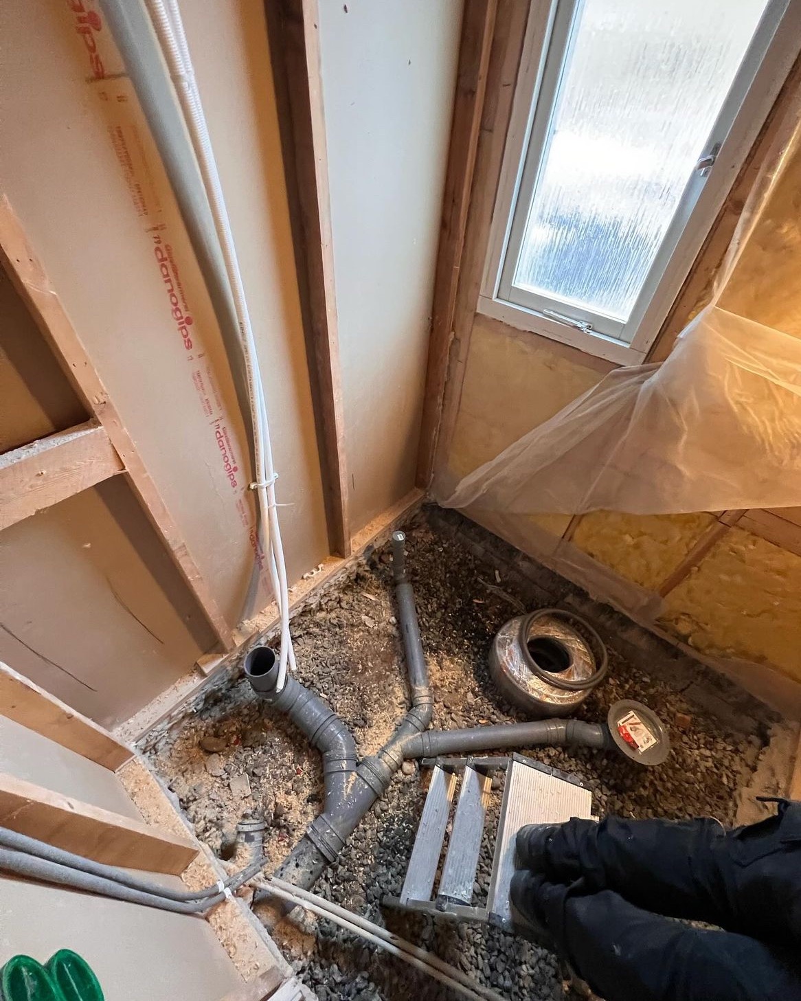 Fragment łazienki w trakcie prac: widoczne rury kanalizacyjne na podłożu z drobnych kamieni, częściowo obudowane ścianami z płyt gipsowo-kartonowych i drewnianą konstrukcją, małe okno w tle.