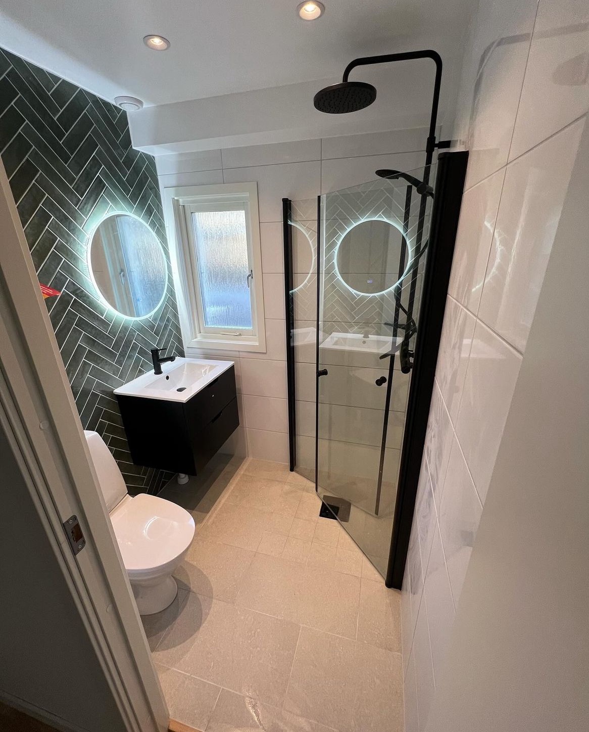 Nowoczesna łazienka z czarnymi akcentami: kabina prysznicowa ze szklanymi drzwiami, umywalka na szafce, toaleta i podświetlane okrągłe lustro na ścianie z ciemnej, ułożonej w jodełkę glazury.