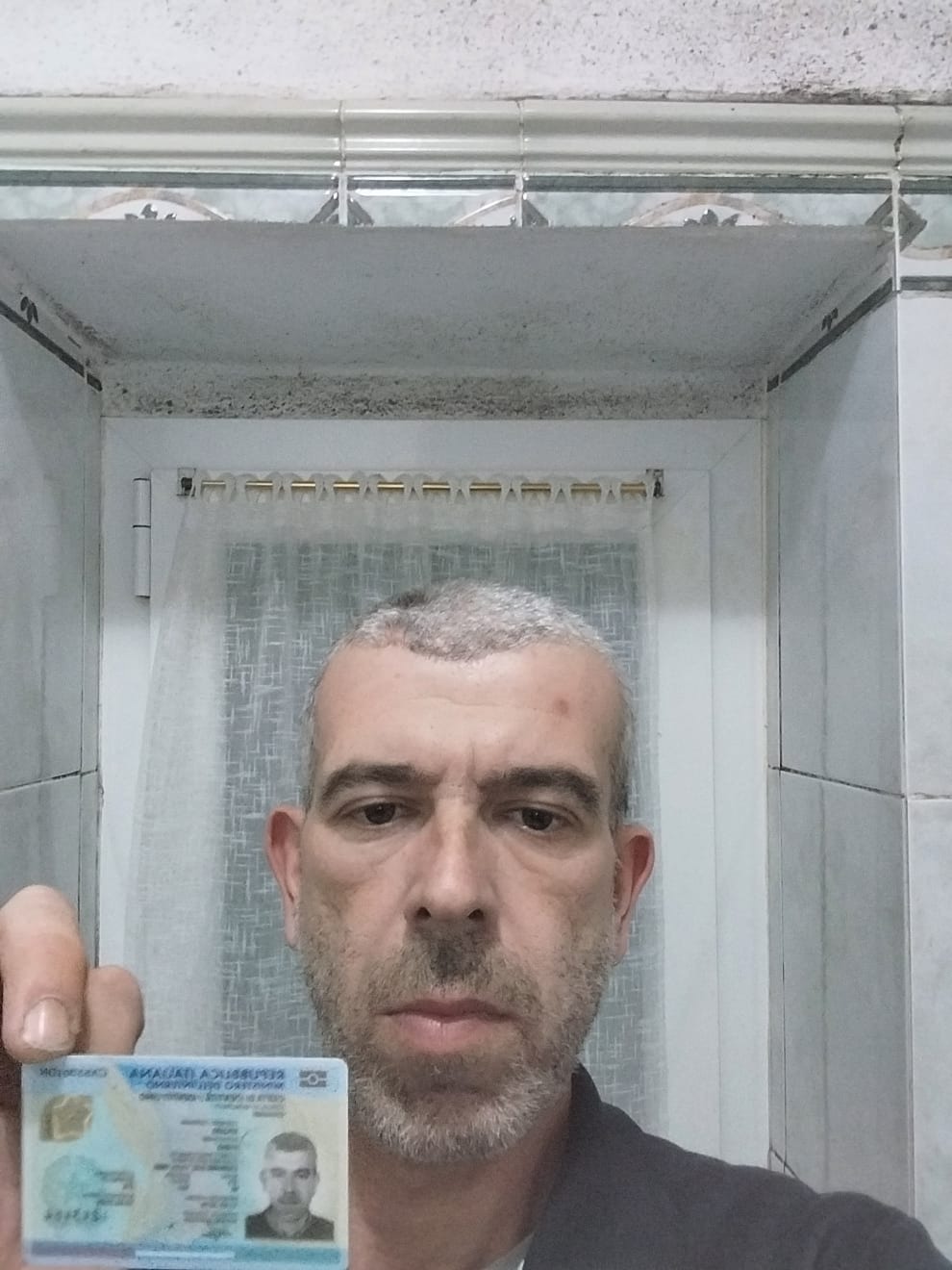Mężczyzna trzymający dowód osobisty na tle łazienki z widoczną pleśnią na suficie.