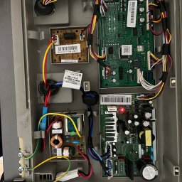 Igor Kupriianow - Wnętrze obudowy lodówki z widocznymi płytkami drukowanymi, przewodami i elementami elektronicznymi. Czerwona dioda LED świeci na jednej z płytek. Widoczne naklejki z kodami kreskowymi...