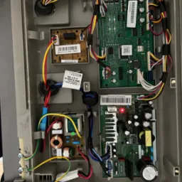 Wnętrze obudowy lodówki z widocznymi płytkami drukowanymi, przewodami i elementami elektronicznymi. Czerwona dioda LED świeci na jednej z płytek. Widoczne naklejki z kodami kreskowymi...