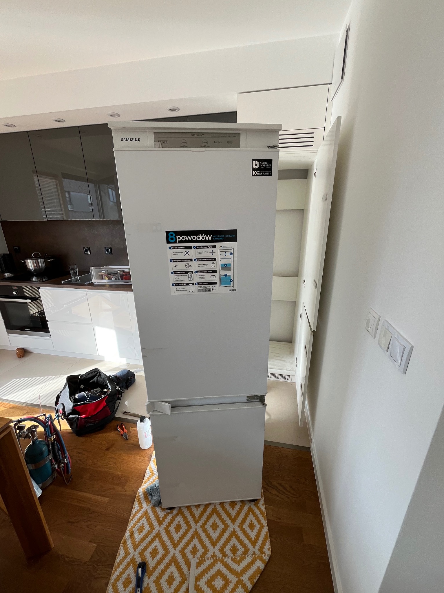 Biała lodówka Samsung w nowoczesnej kuchni podczas naprawy; widoczny otwarty schowek obok, torba narzędziowa, butla z gazem i narzędzia na podłodze