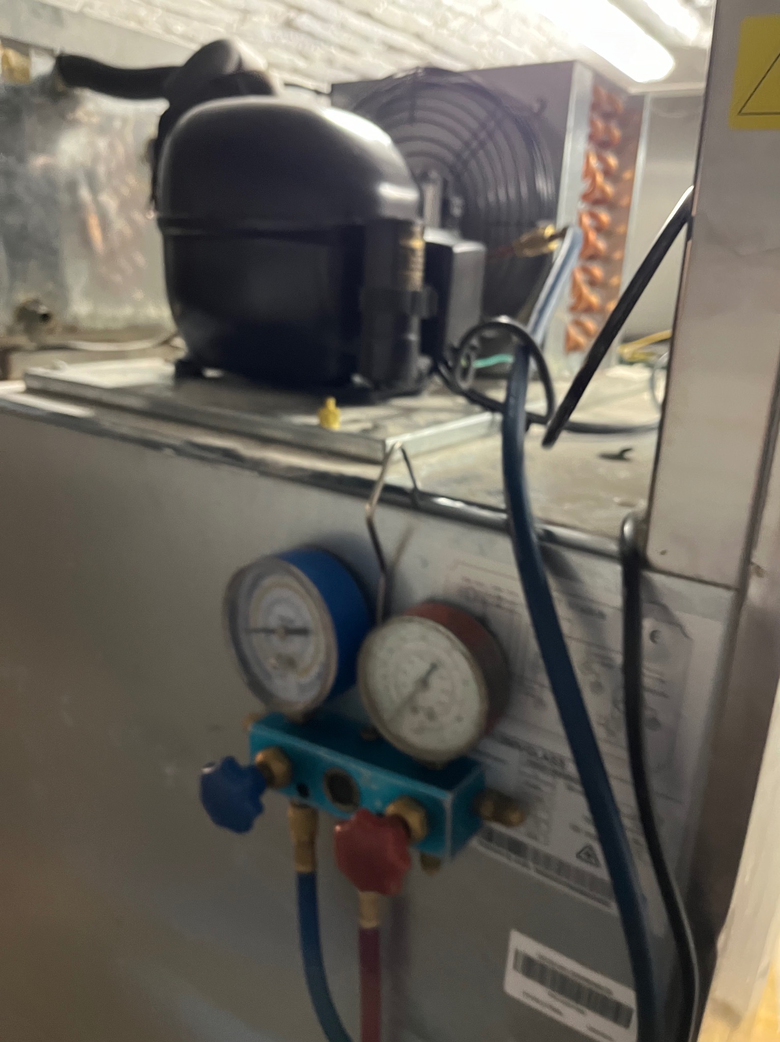 Agregat chłodniczy z podłączonymi manometrami serwisowymi, widoczny wentylator skraplacza, słabe oświetlenie.