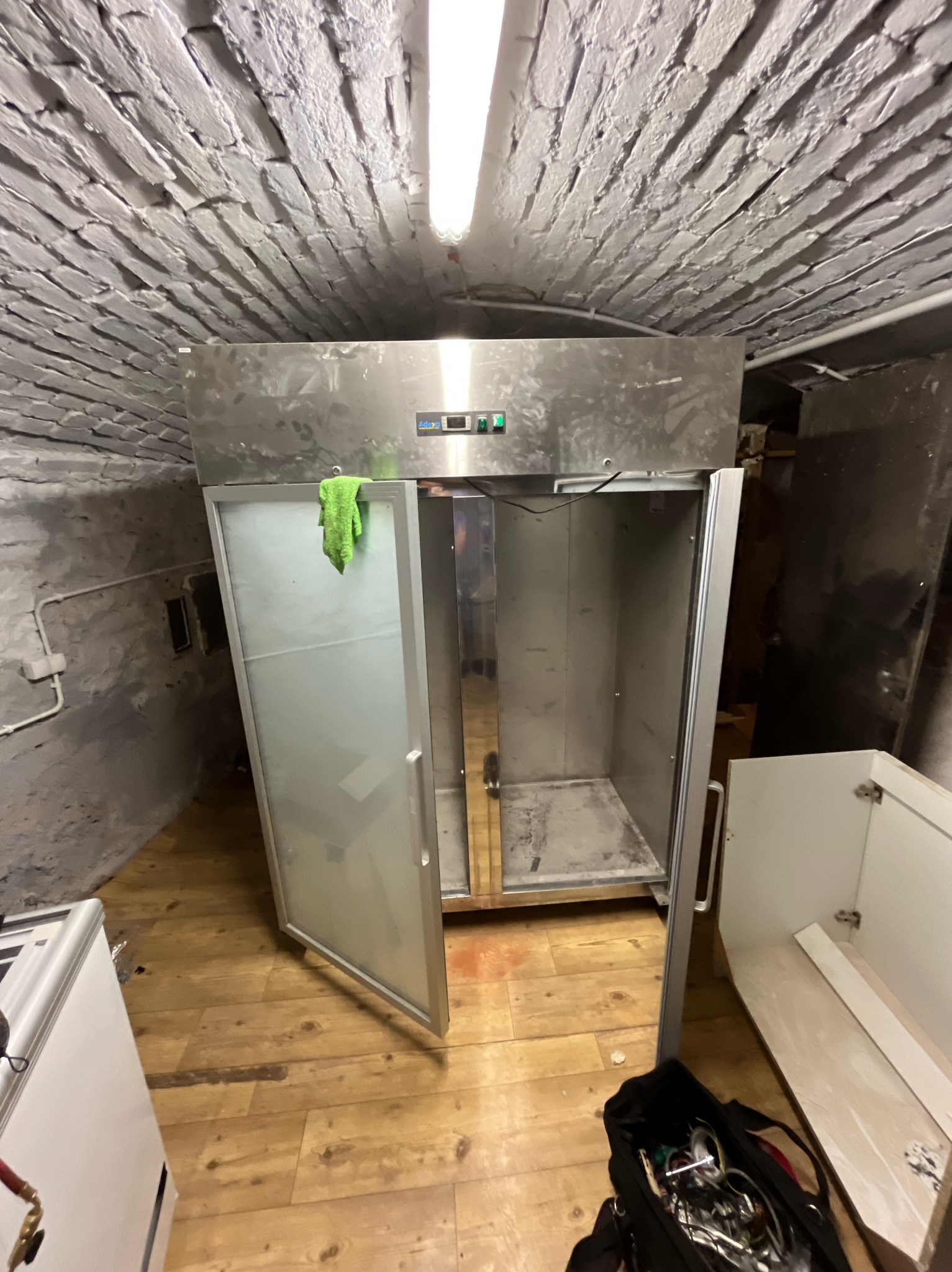 Otwarta, pusta chłodziarka przemysłowa ze stali nierdzewnej w pomieszczeniu z ceglanym sufitem, na drzwiach wisi zielona ściereczka, obok torba z narzędziami.