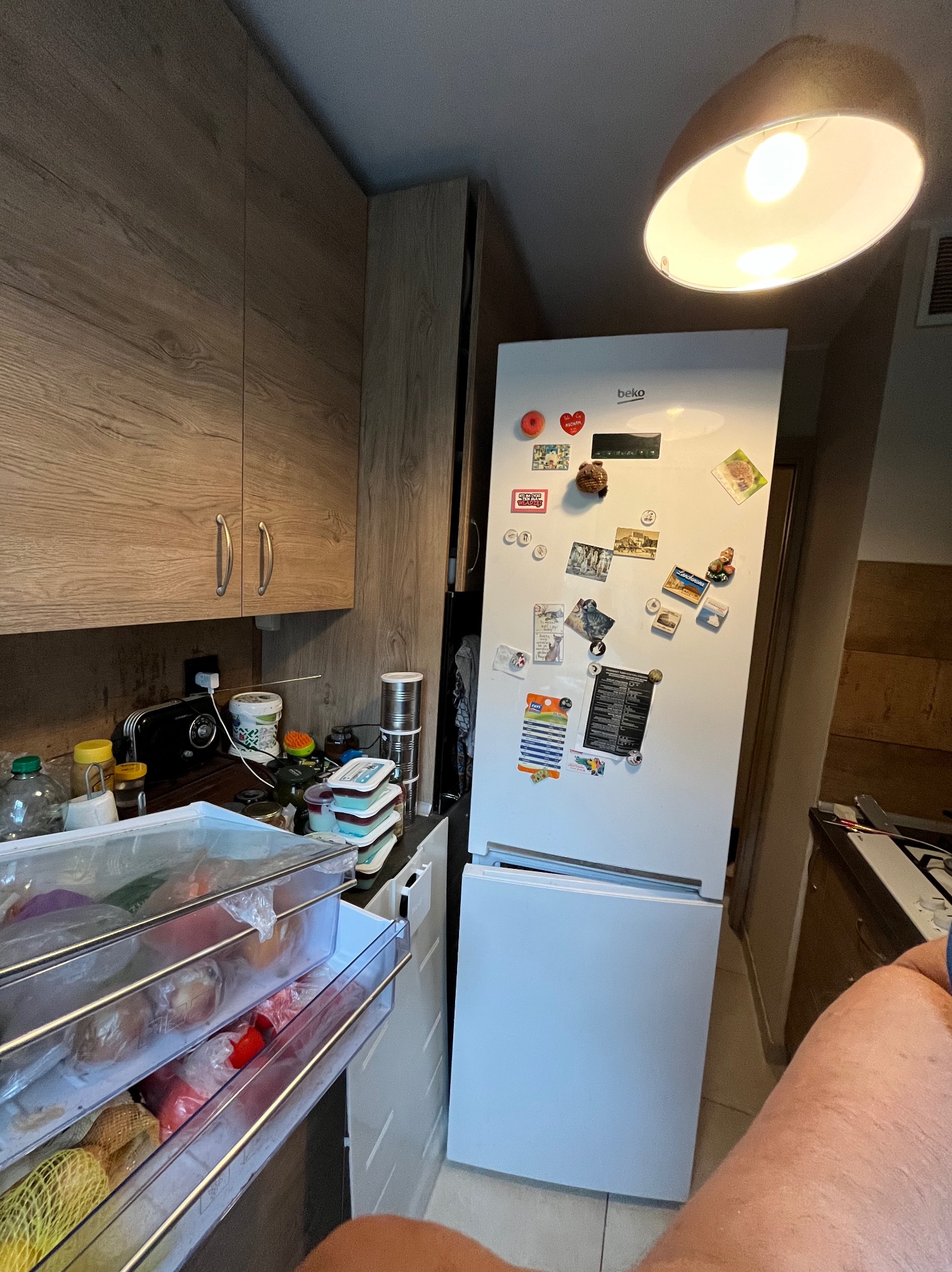 Wnętrze kuchni z białą lodówką Beko, na której przyczepione są magnesy i kartki, wysunięte szuflady lodówki wypełnione produktami spożywczymi, widoczne szafki kuchenne z drewnopodobnymi frontami.
