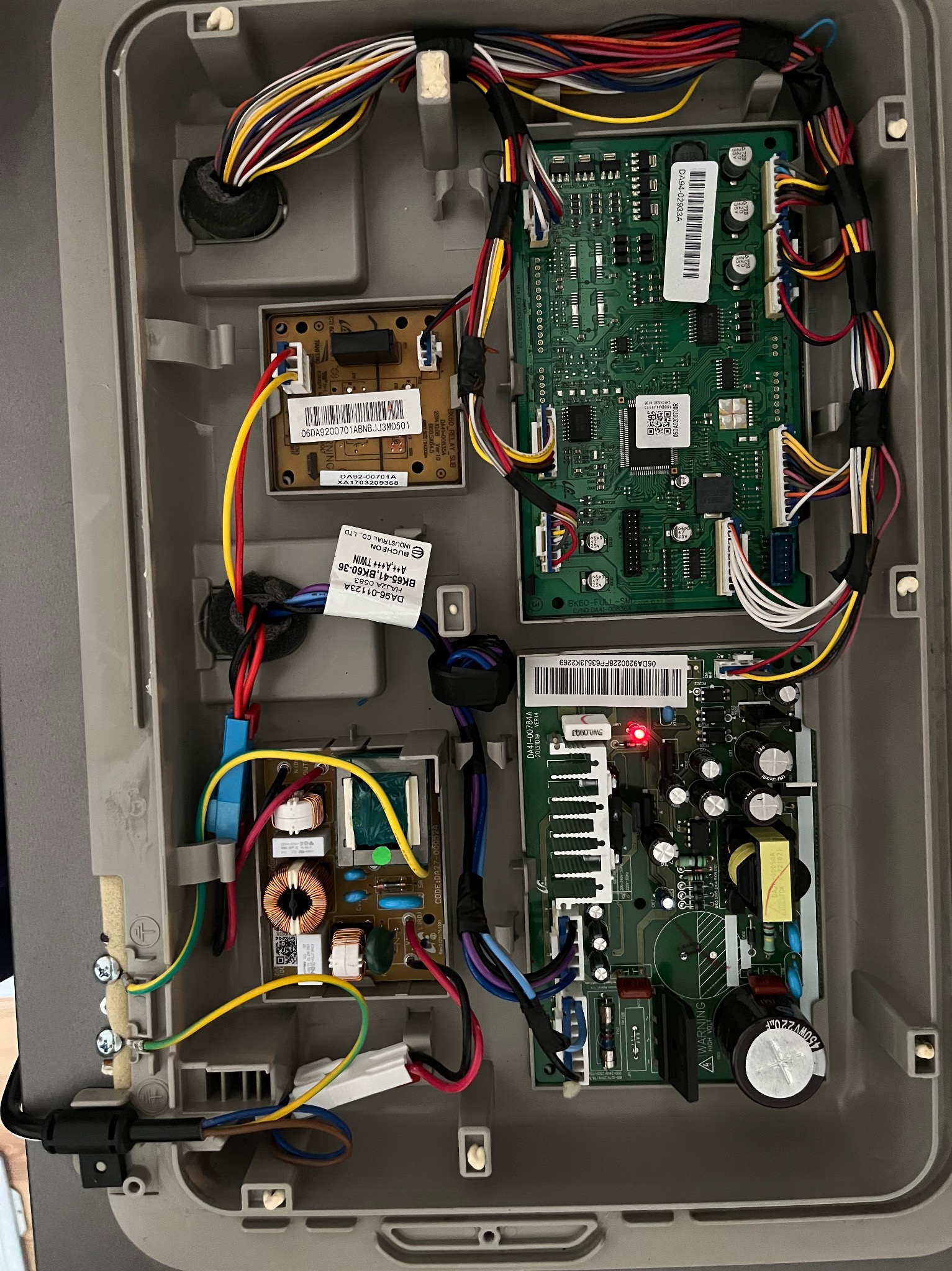 Wnętrze obudowy lodówki z widocznymi płytkami drukowanymi, przewodami i elementami elektronicznymi. Czerwona dioda LED świeci na jednej z płytek. Widoczne naklejki z kodami kreskowymi...