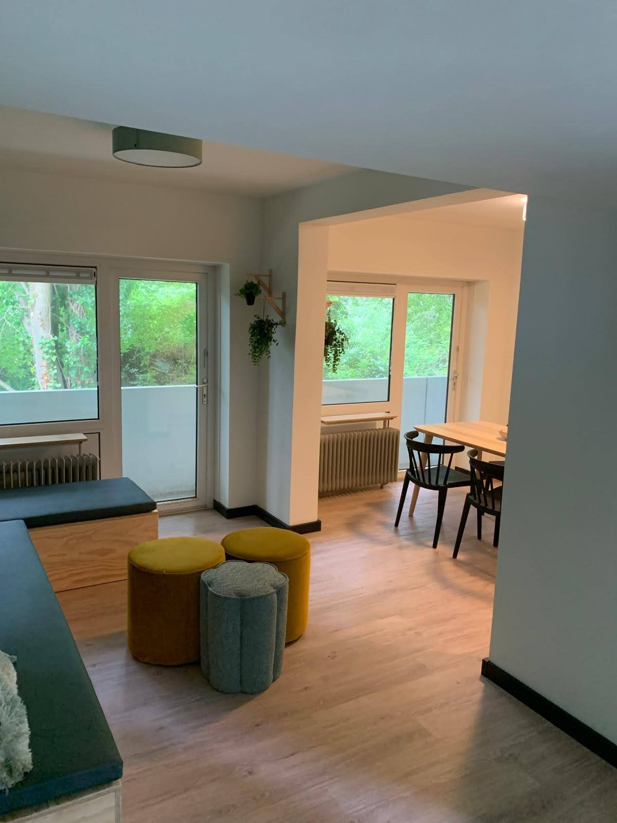 Jasne wnętrze mieszkania z parkietem, widok na balkon z zielenią, siedziska w kolorach żółtym i szarym, oraz fragment jadalni z czarnymi krzesłami.