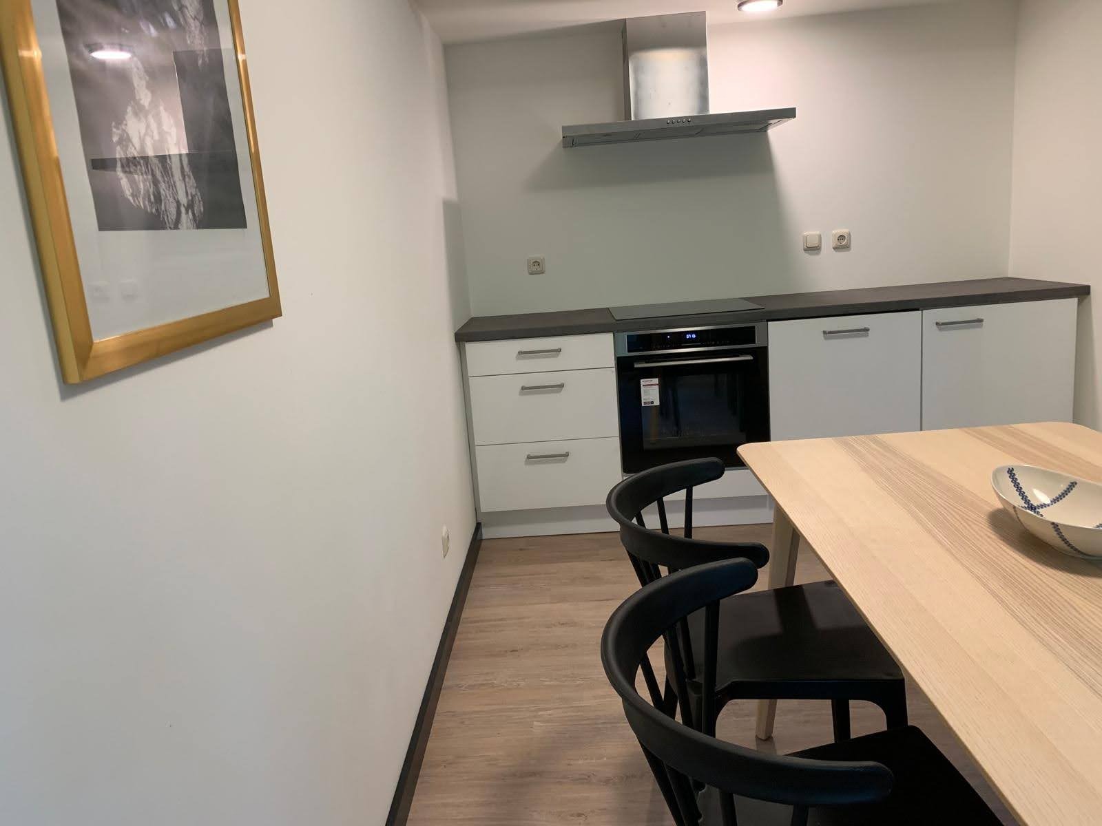 Jasna kuchnia z białymi szafkami, okapem i piekarnikiem, uzupełniona drewnianym stołem i czarnymi krzesłami, z obrazem w złotej ramie na ścianie.