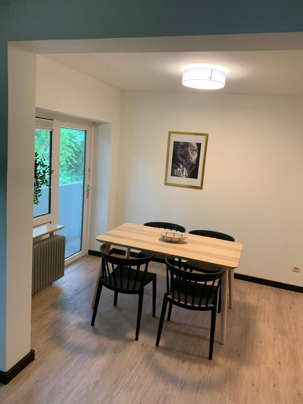 Jasne wnętrze po odnowieniu: drewniany stół z czterema czarnymi krzesłami, obraz w złotej ramie na białej ścianie, widok na zieleń za oknem.