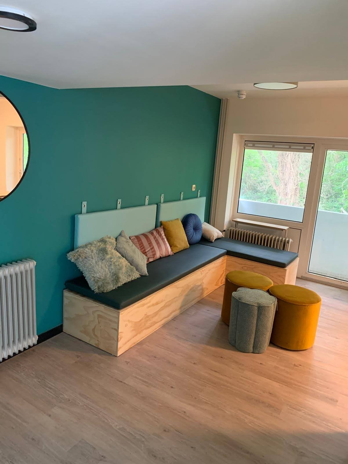 Turkusowa ściana w pokoju z drewnianą zabudową siedziska z poduszkami, obok okna grzejnik i pufy w odcieniach żółci i szarości.