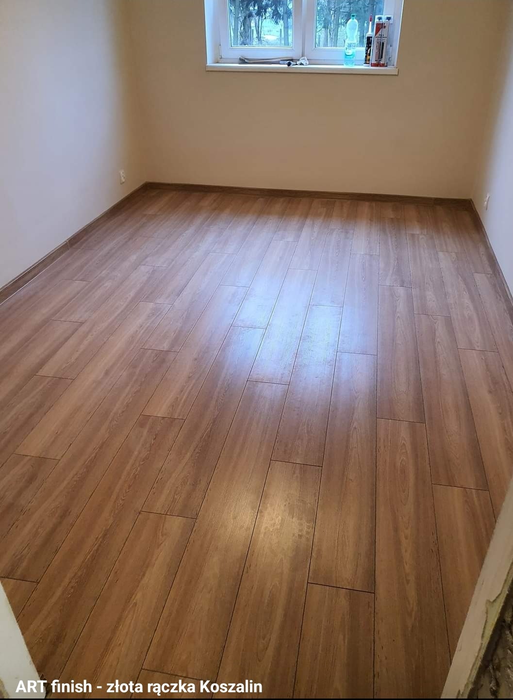 Wykończone pomieszczenie z nową, brązową podłogą z paneli imitujących drewno, widok od progu, ściany w odcieniach beżu, okno z widokiem na drzewa.