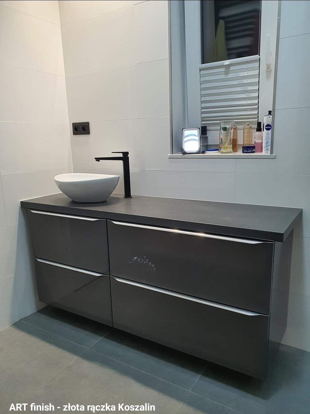 Nowoczesna łazienka z szarą szafką pod umywalkę nablatową i czarną armaturą, białe płytki na ścianach i szare na podłodze, okno z żaluzjami w tle.