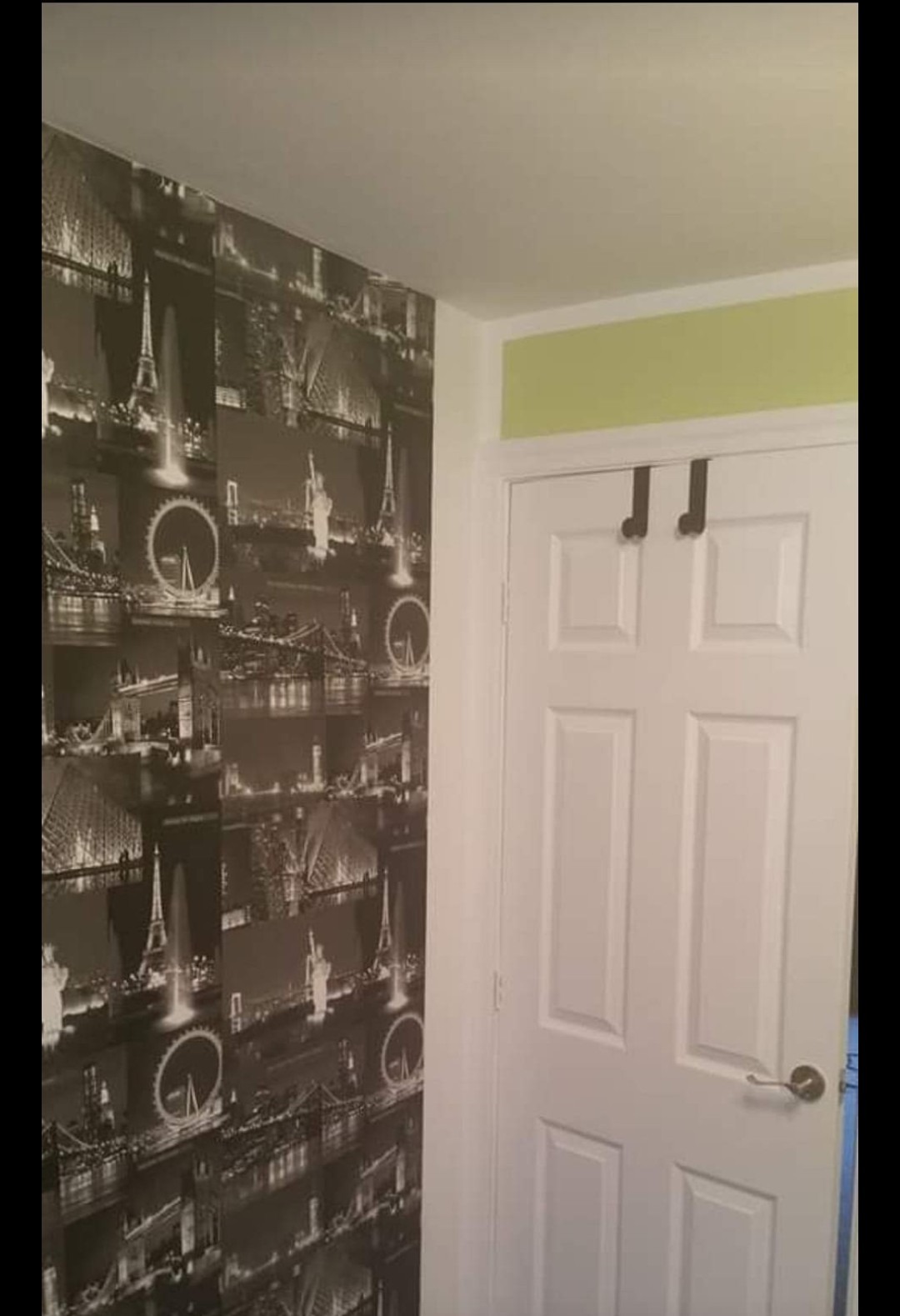 Ściana z tapetą w odcieniach szarości przedstawiającą panoramy miast, widoczna obok białych drzwi z haczykami na górze i fragmentem ściany pomalowanej na jasnozielono.