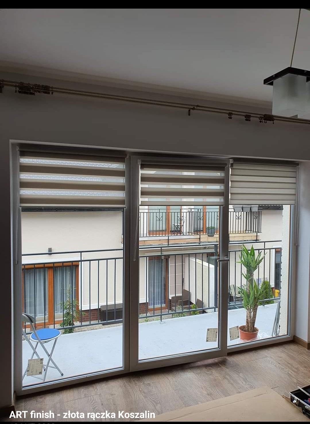 Zasłonięte okna trzyskrzydłowe z widokiem na balkon w bloku z rośliną w doniczce i niebieskim krzesłem składanym, zamontowane karnisze na ścianie.