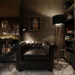 Anna Bartman - Z WNĘTRZA - Elegancki, ciemny salon w Krakowie z skórzanym fotelem Chesterfield, drewnianym regałem z książkami, stolikiem kawowym z butelkami alkoholu i dekoracyjnymi maskami na ścianie.