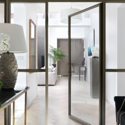 Anna Bartman - Z WNĘTRZA - Nowoczesne wnętrze mieszkania z otwartymi szklanymi drzwiami, przez które widać salon z sofą i roślinami, na pierwszym planie stolik z ozdobną lampą i orchideami oraz czarny aksamitny fotel z torebką.