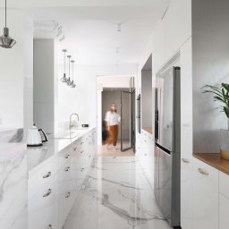 Anna Bartman - Z WNĘTRZA - Wnętrze nowoczesnej kuchni z białymi szafkami, marmurowym blatem i podłogą, oświetleniem punktowym i wiszącymi lampami, z widoczną osobą w tle.