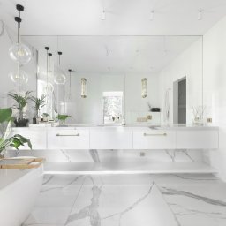 Anna Bartman - Z WNĘTRZA - Jasna łazienka z marmurową podłogą i ścianami, biała wanna z złotą armaturą i roślinami, duże lustro nad podwieszaną szafką z umywalkami.