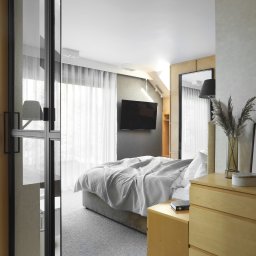 Anna Bartman - Z WNĘTRZA - Nowoczesna sypialnia z łóżkiem przykrytym szarą kołdrą, drewnianymi szafkami nocnymi, czarnym telewizorem na ścianie i szklanymi drzwiami z czarną ramą.