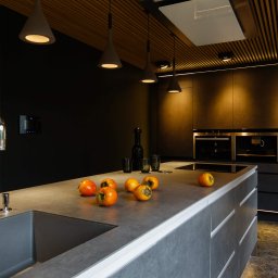 Anna Bartman - Z WNĘTRZA - Nowoczesna kuchnia w ciemnych barwach z wyspą, oświetleniem punktowym i owocami persymony na blacie.