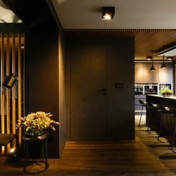 Anna Bartman - Z WNĘTRZA - Nowoczesne wnętrze: ciemna kuchnia z wyspą i hokerami, drewniany sufit lamelowy, podłoga z ciemnego drewna, minimalistyczne meble i dekoracje, lampa podłogowa i kwiaty na stoliku.