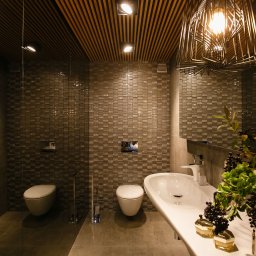 Anna Bartman - Z WNĘTRZA - Nowoczesna łazienka z mozaikową ścianą, podwieszaną toaletą, umywalką na blacie, dekoracyjnym oświetleniem i drewnianym sufitem listwowym. Na blacie umywalki dekoracja z roślin i kosmetyków.