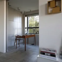 Jasne wnętrze z drewnianym biurkiem i krzesłem przy oknie, minimalistyczna dekoracja ścienna, książki i szklane wazony na białej komodzie, w tle przeszklona witryna z alkoholem.