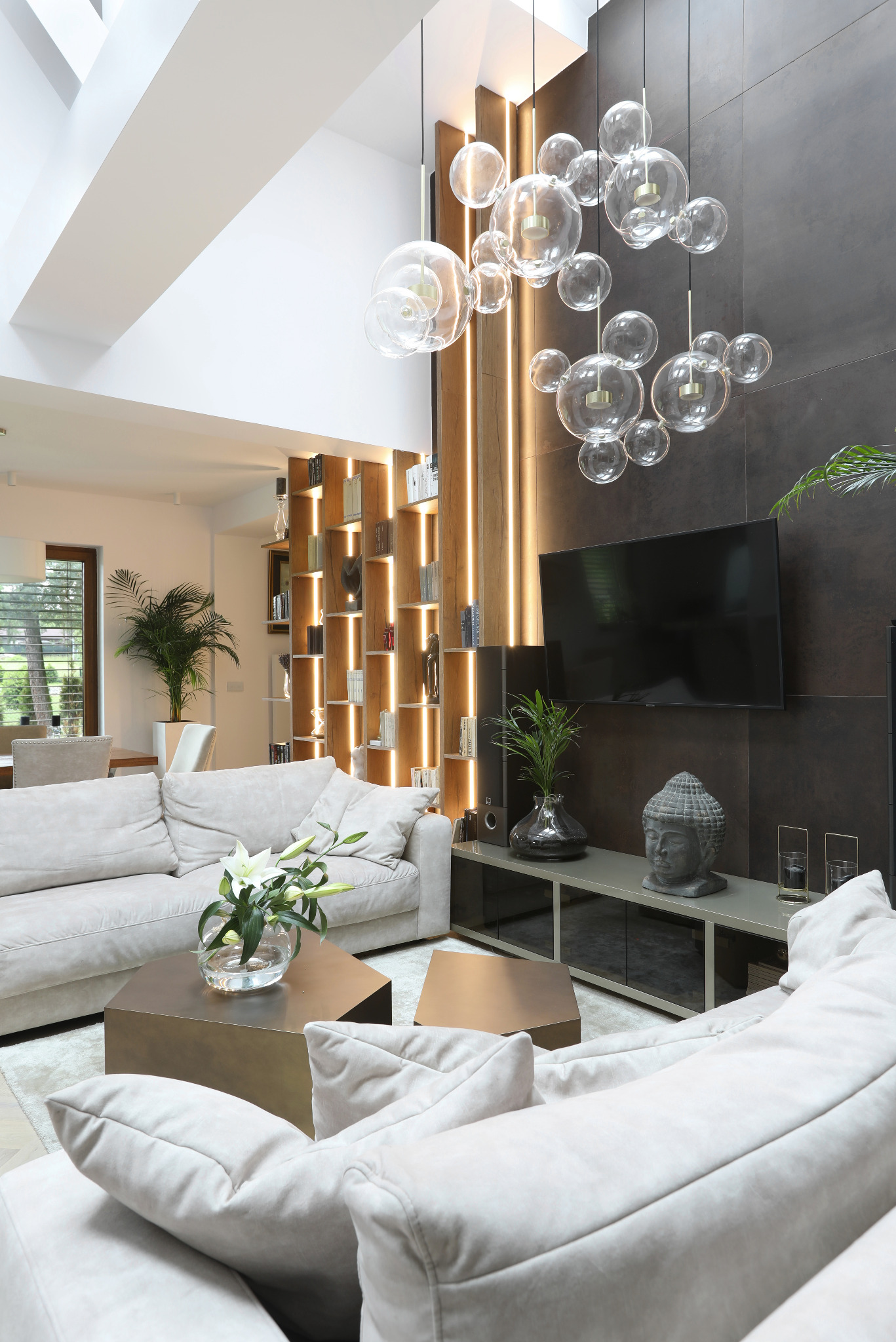 Jasny salon z wysokim sufitem, szarobeżowa sofa, stoliki kawowe w kolorze mosiądzu, nowoczesna lampa z szklanych kul, podświetlany regał na książki i ciemna ściana z telewizorem
