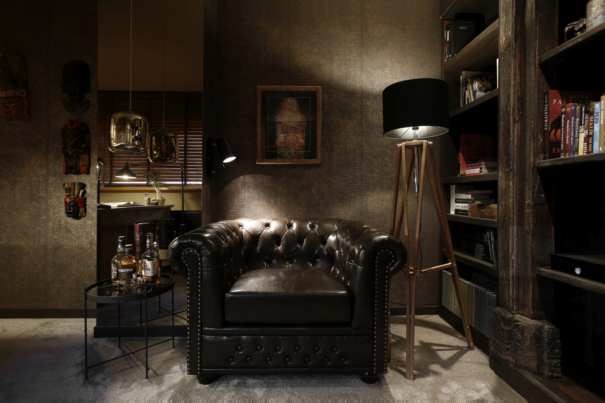 Ciemne wnętrze pokoju z skórzanym fotelem typu chesterfield, stolikiem z butelkami alkoholu, lampą stojącą i regałem z książkami, utrzymane w stylu retro.
