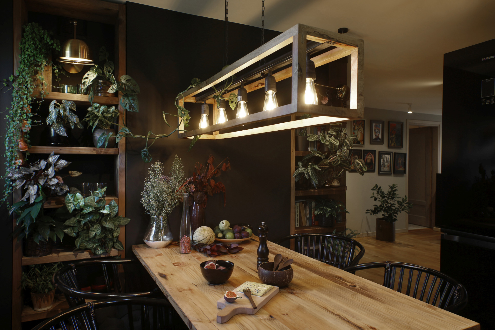 Rustykalny stół z jasnego drewna, nakryty owocami, serem i figami, oświetlony wiszącą lampą w drewnianej ramie, z roślinami pnącymi się po półkach w tle, w pomieszczeniu z czarną ścianą i widoczną...