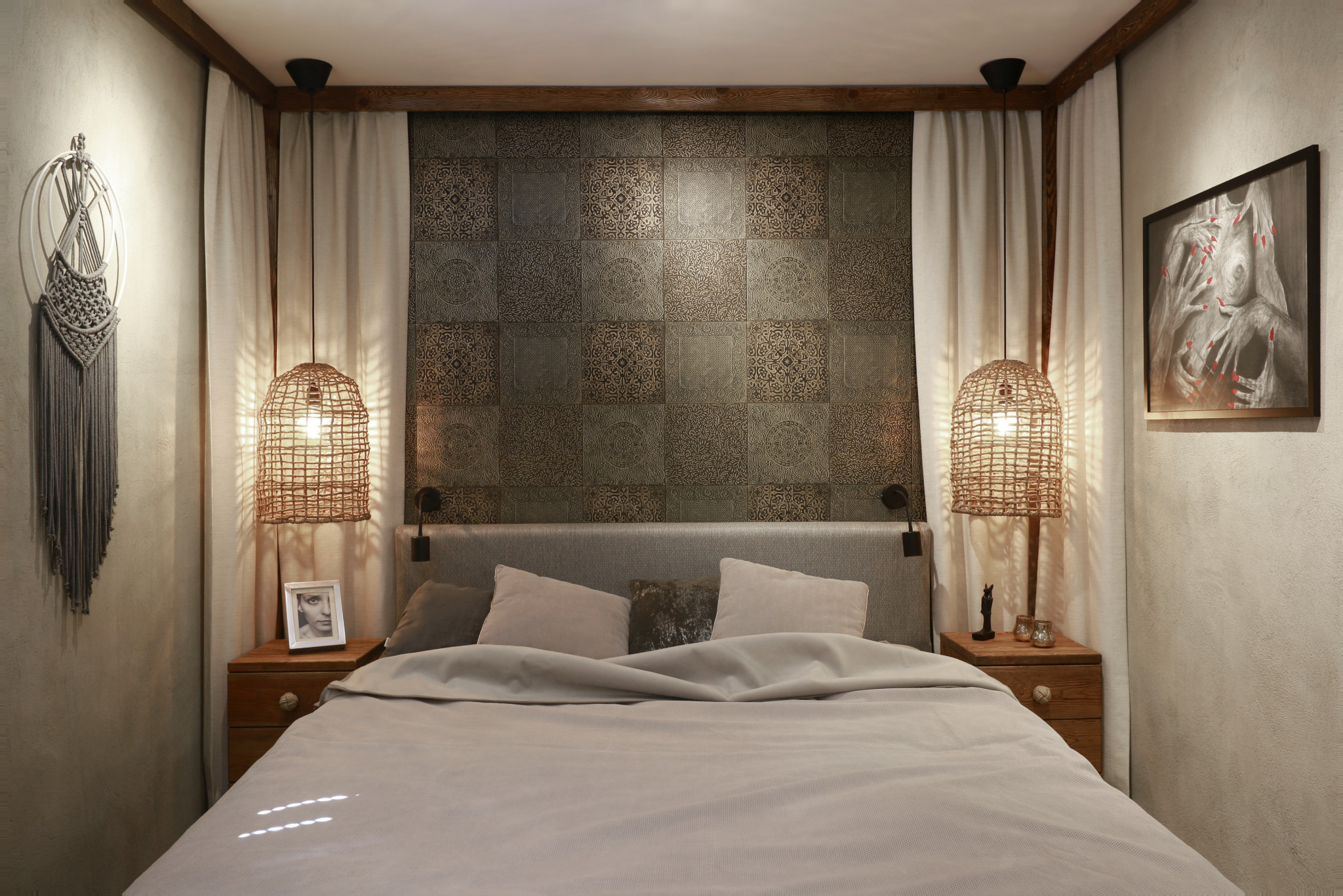 Sypialnia w stylu boho z łóżkiem z szarą ramą, wzorzystą tapetą za wezgłowiem, lampami z abażurami z wikliny, makramą na ścianie i drewnianymi szafkami nocnymi z dekoracjami.