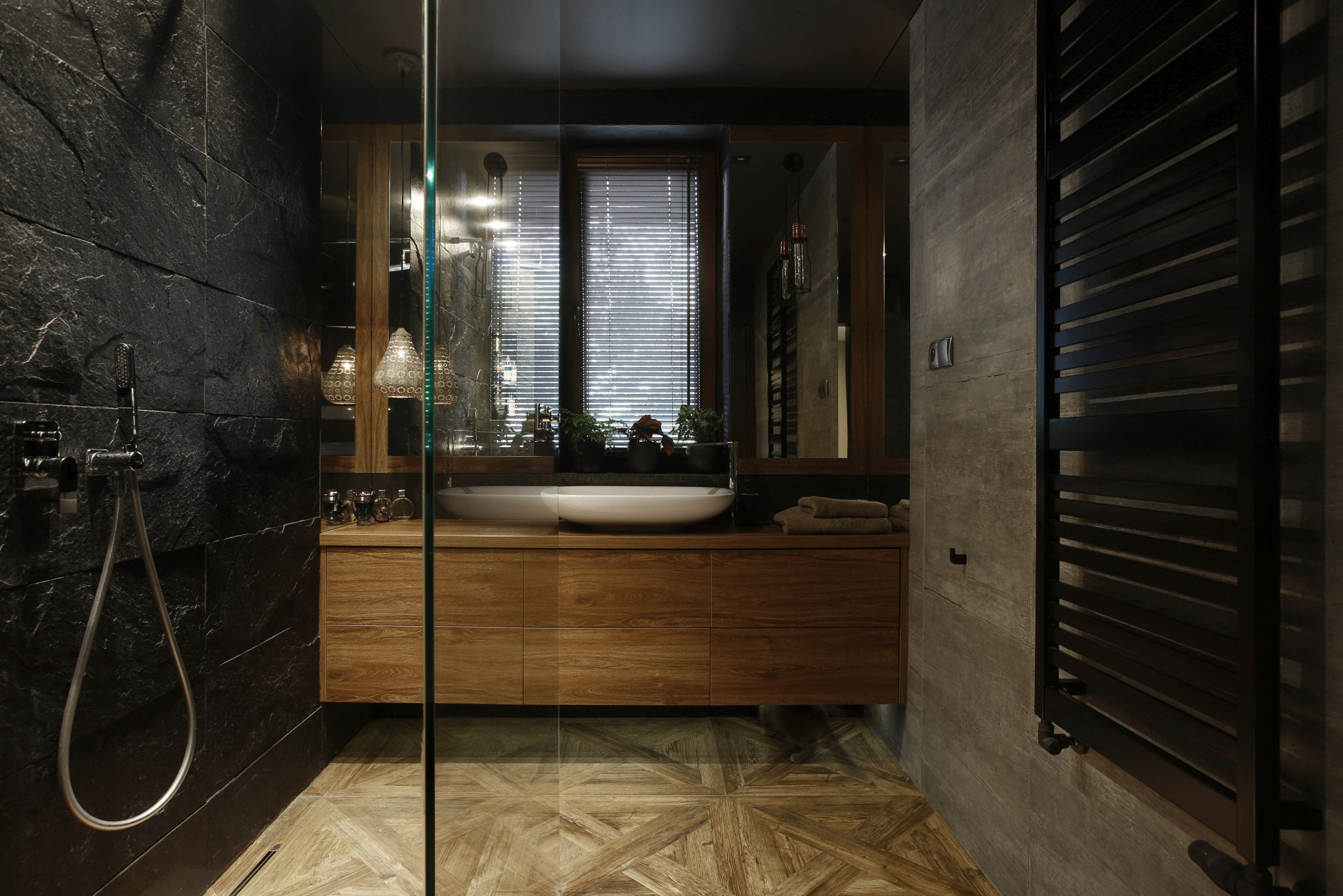 Nowoczesna łazienka z czarnymi płytkami imitującymi łupek na ścianie prysznicowej, drewnianą szafką pod umywalką z dwiema białymi umywalkami nablatowymi, grzejnikiem drabinkowym w kolorze...