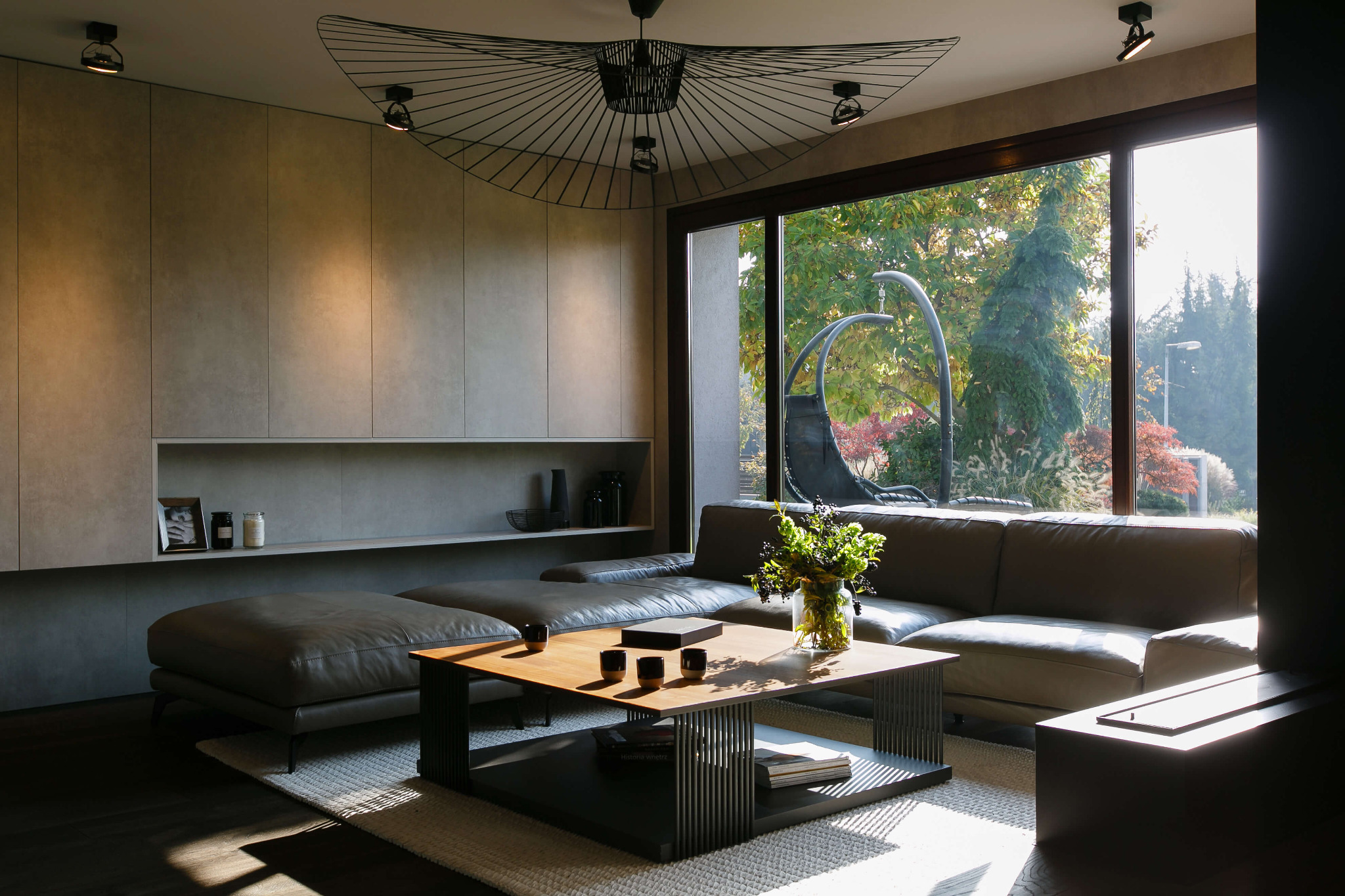 Nowoczesny salon z dużym oknem, skórzaną sofą narożną, stolikiem kawowym z drewnianym blatem i metalową podstawą oraz dekoracyjną lampą sufitową w stylu pająka. Za oknem widoczny ogród z huśtawką.