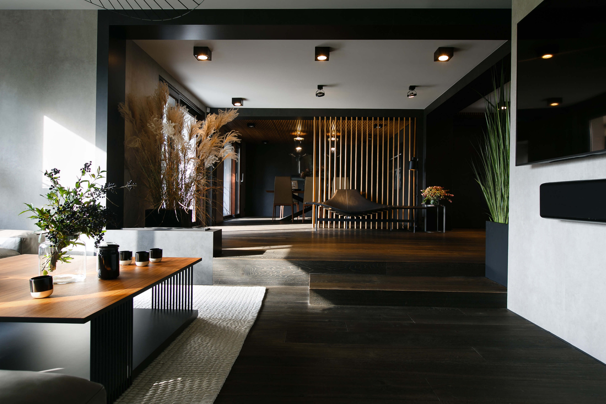 Przestronne wnętrze domu w ciemnej tonacji z drewnianą podłogą, stołem z dekoracjami roślinnymi i ażurową ścianką działową z drewna.
