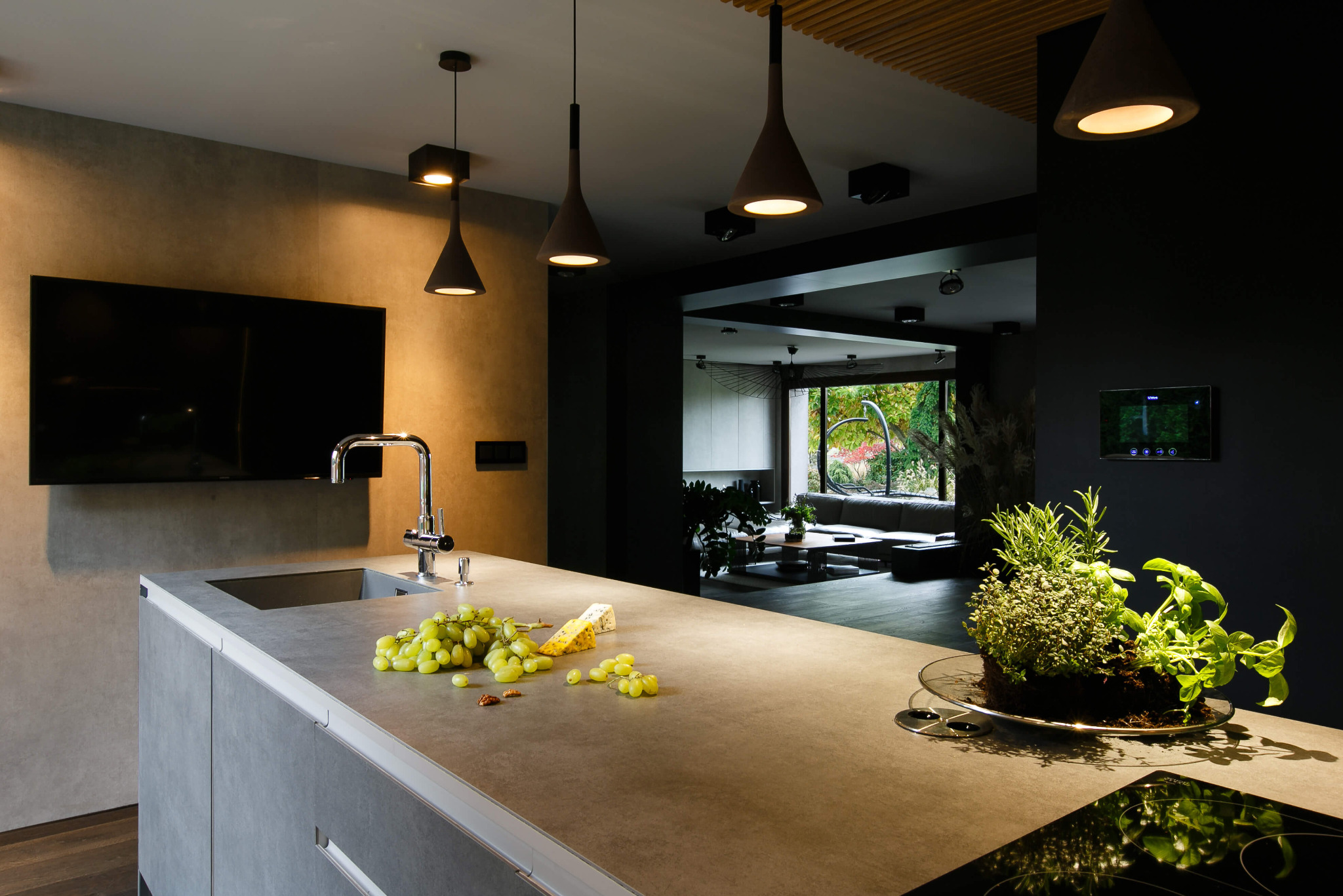 Nowoczesna kuchnia z szarą wyspą, na której leżą winogrona, ser i zioła w doniczce; w tle widoczny salon z dużym oknem.