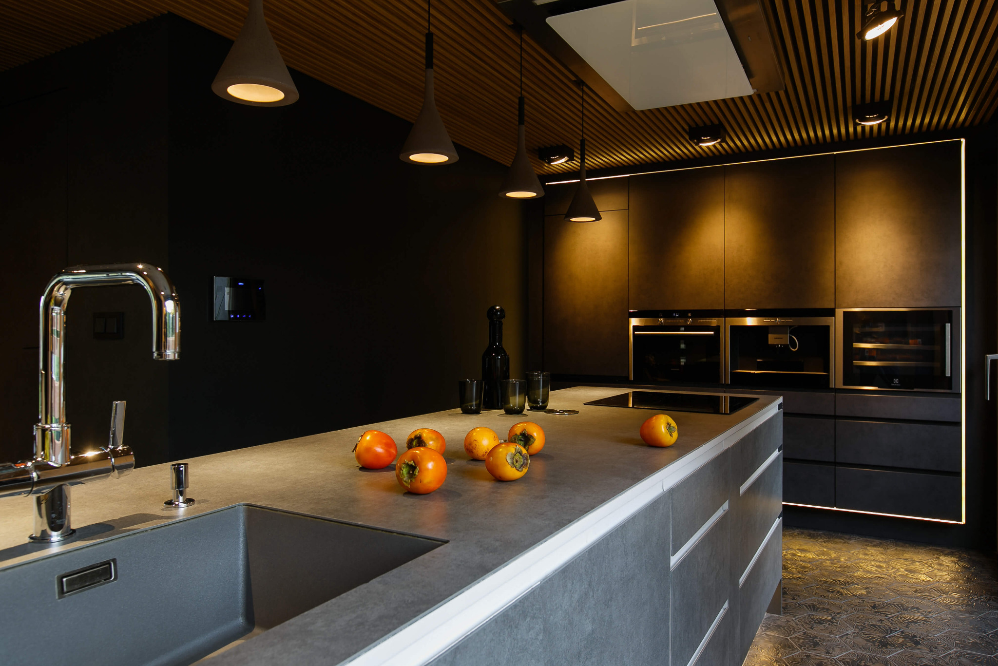Nowoczesna kuchnia w ciemnych barwach z wyspą, oświetleniem punktowym i owocami persymony na blacie.