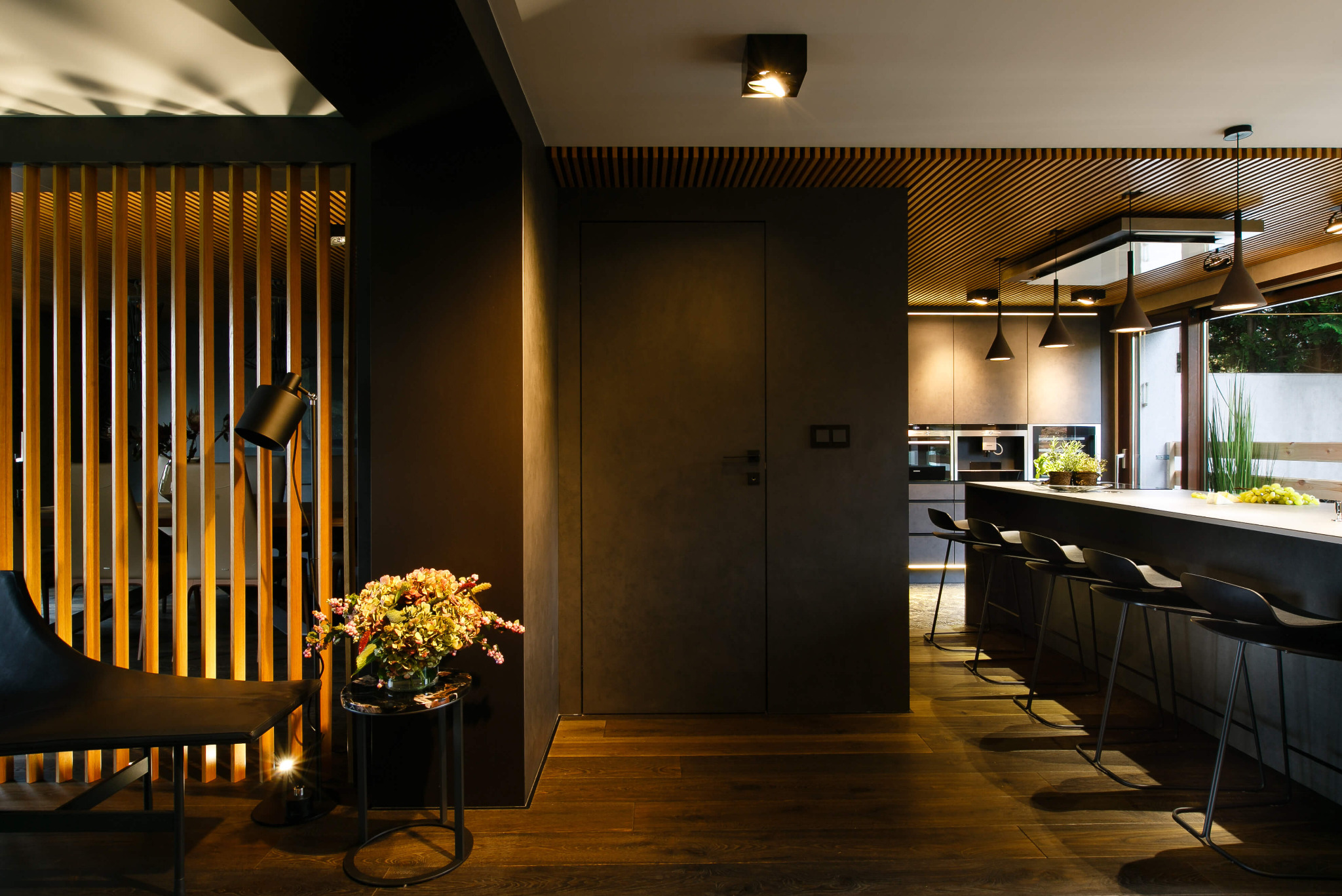 Nowoczesne wnętrze: ciemna kuchnia z wyspą i hokerami, drewniany sufit lamelowy, podłoga z ciemnego drewna, minimalistyczne meble i dekoracje, lampa podłogowa i kwiaty na stoliku.