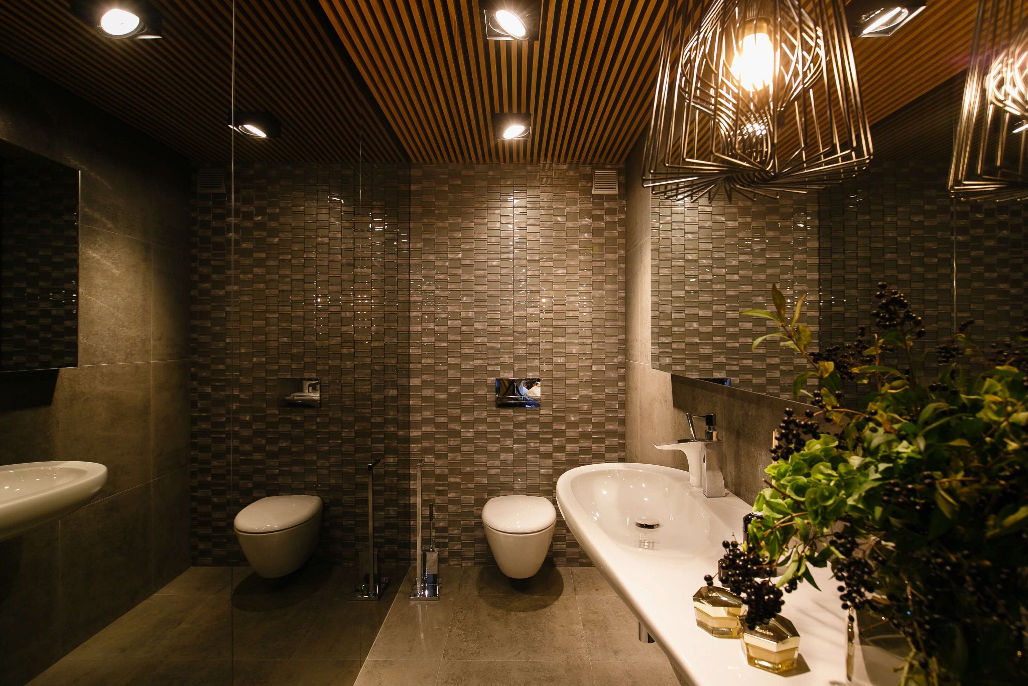 Nowoczesna łazienka z mozaikową ścianą, podwieszaną toaletą, umywalką na blacie, dekoracyjnym oświetleniem i drewnianym sufitem listwowym. Na blacie umywalki dekoracja z roślin i kosmetyków.