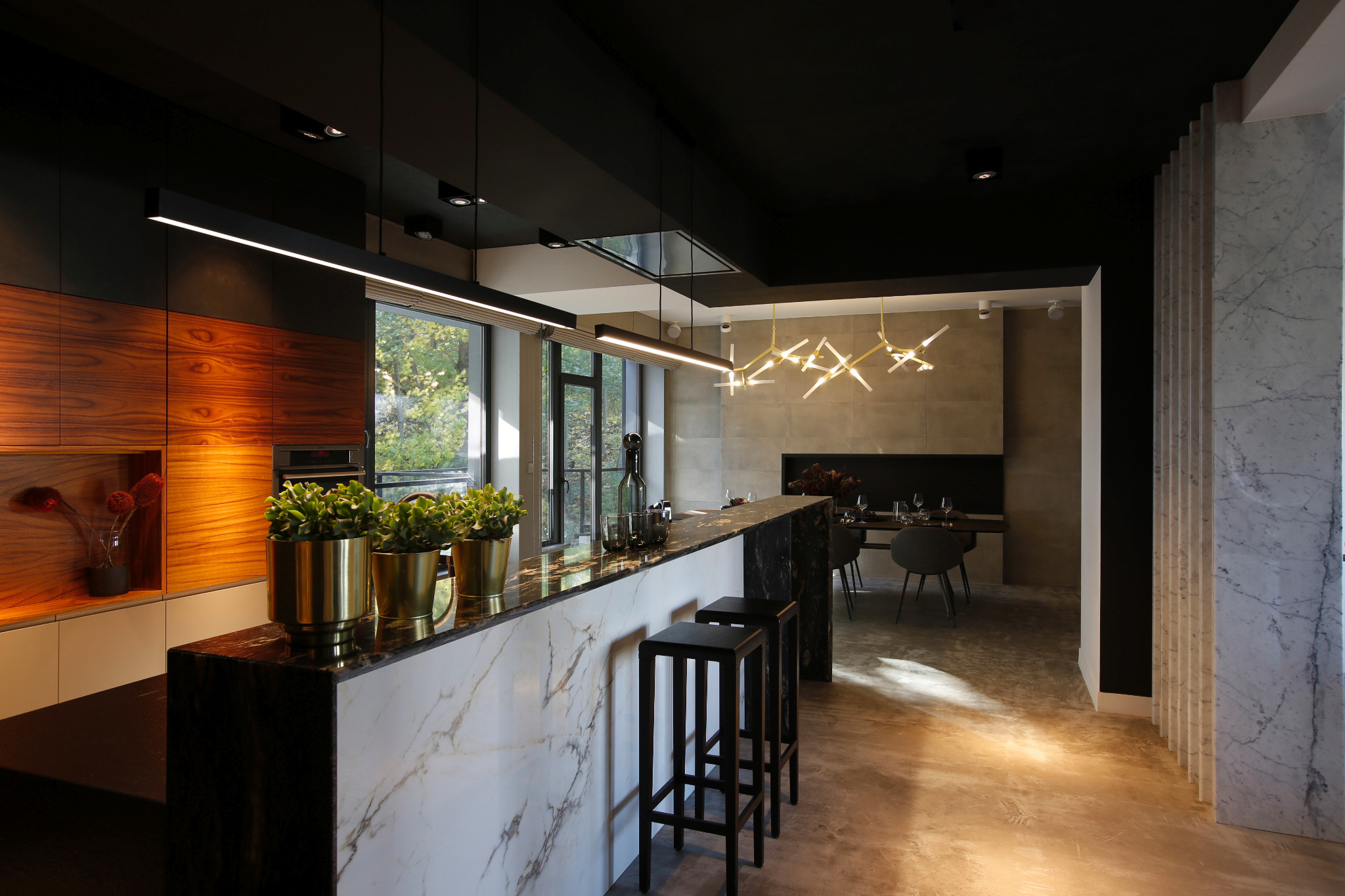 Nowoczesna kuchnia z wyspą wykończoną marmurem, czarnymi stołkami barowymi i złotymi doniczkami z zielonymi roślinami. W tle jadalnia z minimalistycznym oświetleniem i szarymi ścianami.