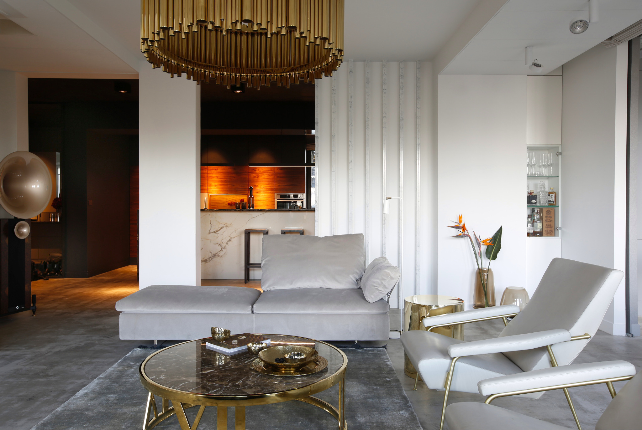Elegancki salon z szarym narożnikiem, marmurowym stolikiem kawowym ze złotymi akcentami, złotym żyrandolem i widoczną kuchnią z drewnianymi frontami i marmurowym blatem w tle.