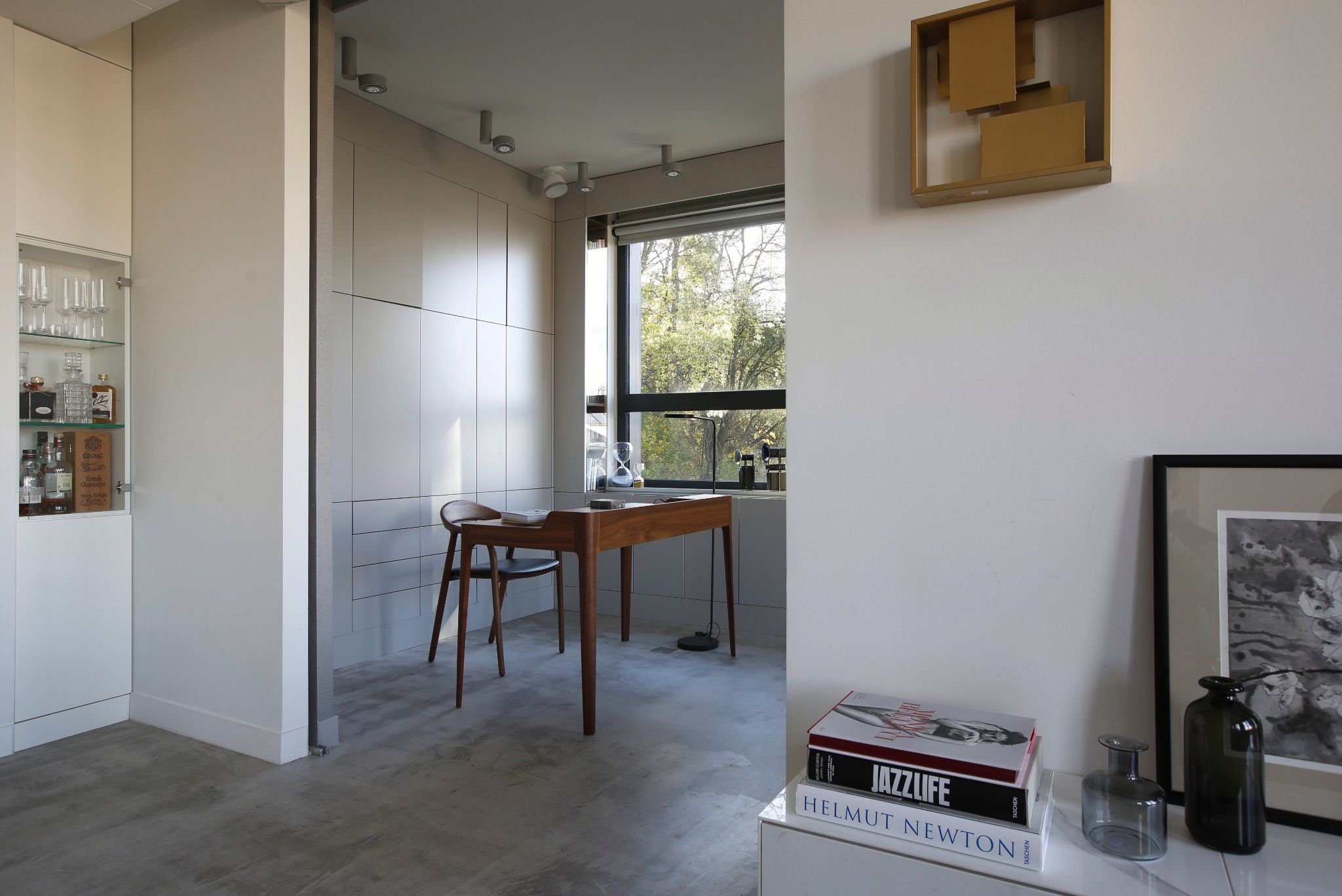 Jasne wnętrze z drewnianym biurkiem i krzesłem przy oknie, minimalistyczna dekoracja ścienna, książki i szklane wazony na białej komodzie, w tle przeszklona witryna z alkoholem.