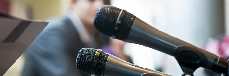 Dwa mikrofony na statywach ustawione na tle rozmytej sylwetki osoby w garniturze i fragmentu dokumentu, sugerujące konferencję lub prezentację.