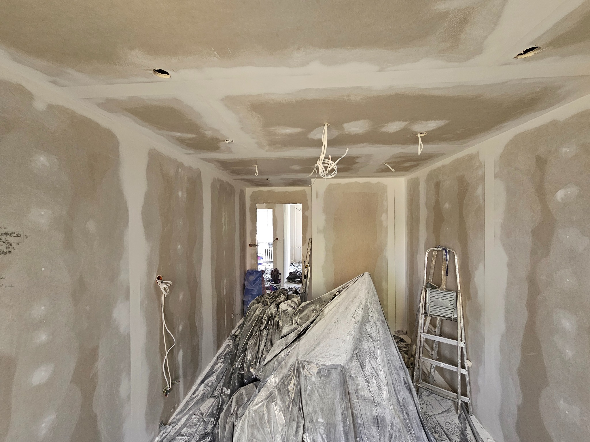 Pokój w trakcie szpachlowania ścian i sufitu, zabezpieczony folią malarską, widoczna drabina i wystające kable. Etap prac wykończeniowych przed malowaniem.