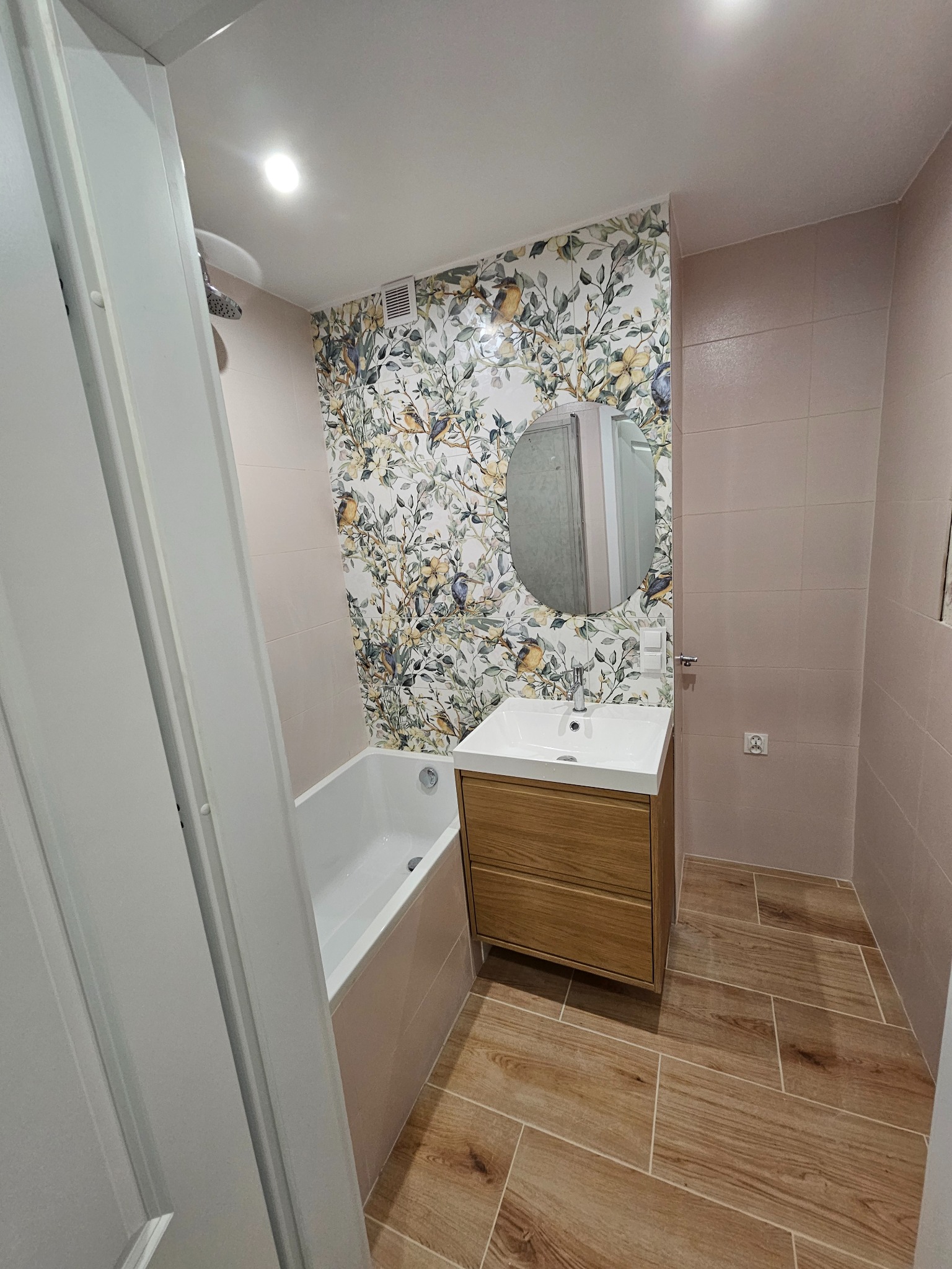 Ukończona łazienka z wanną, umywalką z szafką z drewna i lustrem. Na ścianie płytki z motywem roślinnym i ptaków. Podłoga wyłożona płytkami imitującymi drewno.