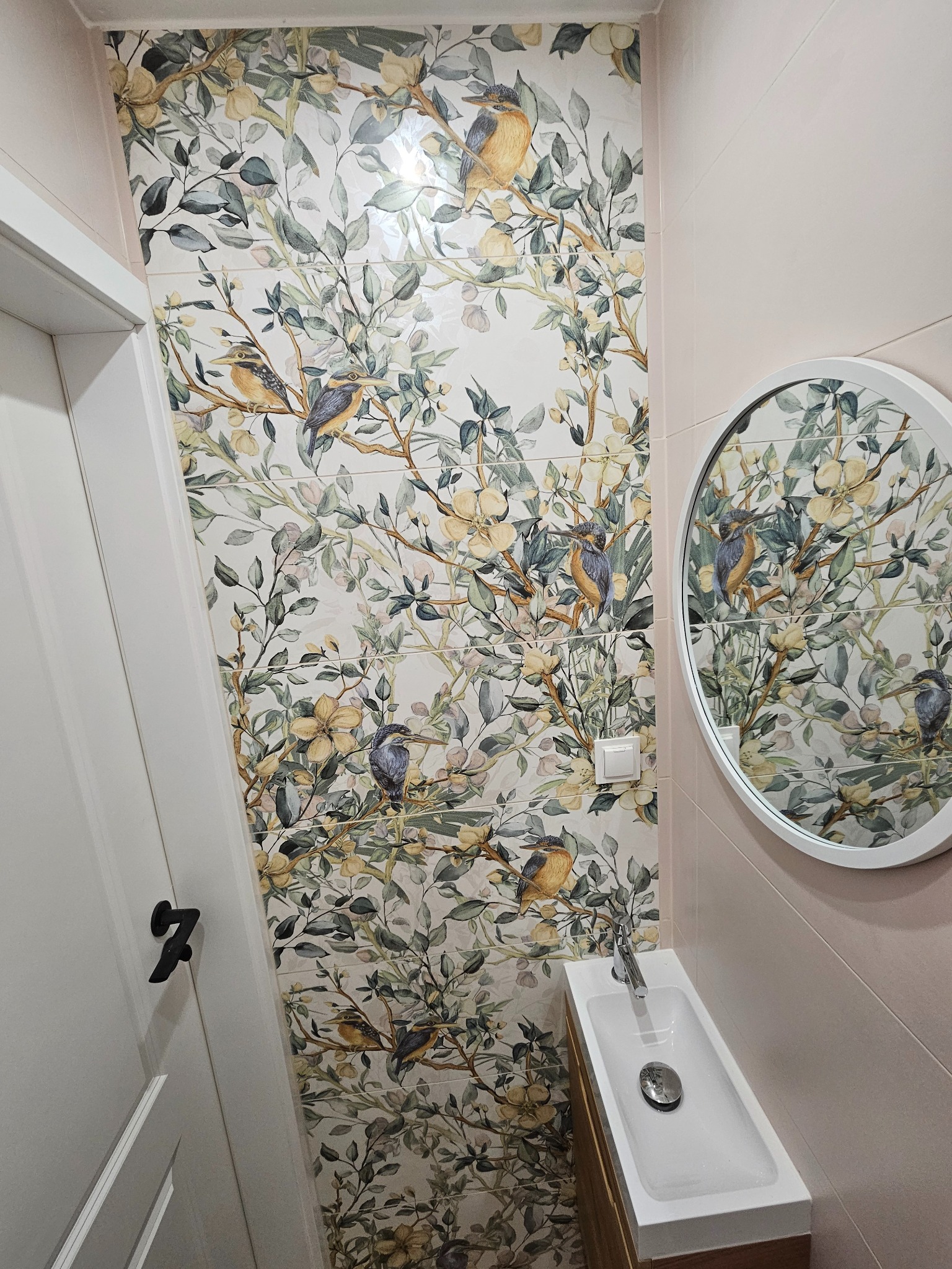 Ukończona łazienka z płytkami w kwiaty i ptaki, okrągłe lustro, wąska umywalka z szafką. Białe drzwi z czarną klamką. Pastelowa kolorystyka.
