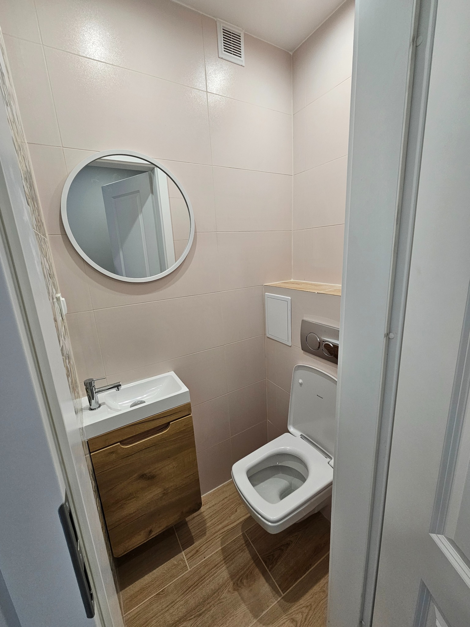 Jasna łazienka z beżowymi płytkami, drewnianą szafką pod umywalką i okrągłym lustrem. Widoczna toaleta i fragment drzwi. Minimalistyczny design.