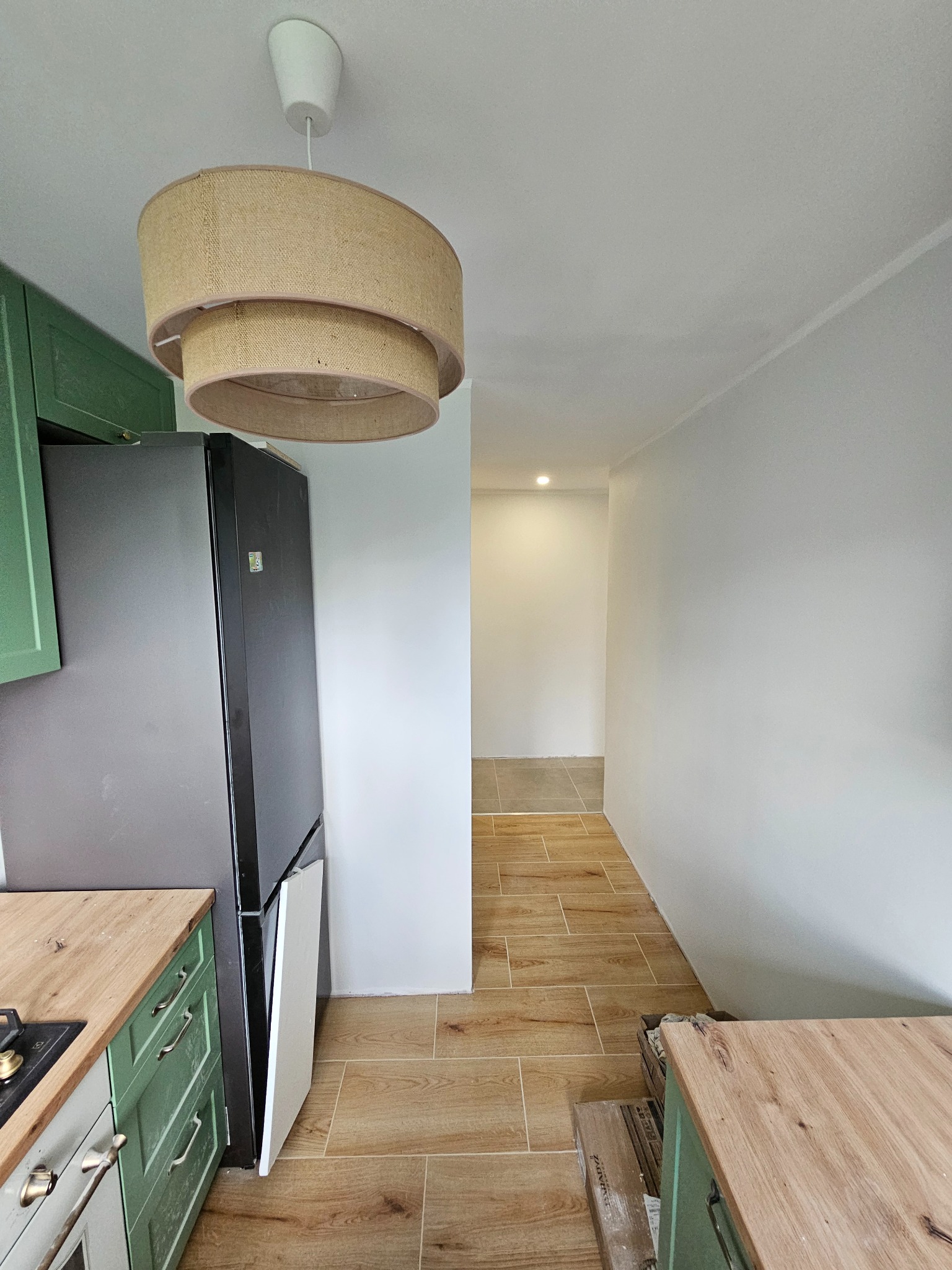 Kuchnia po remoncie: zielone szafki, drewniany blat, brązowe płytki podłogowe imitujące drewno, nowoczesna lampa. Widok na korytarz z jasnymi ścianami.