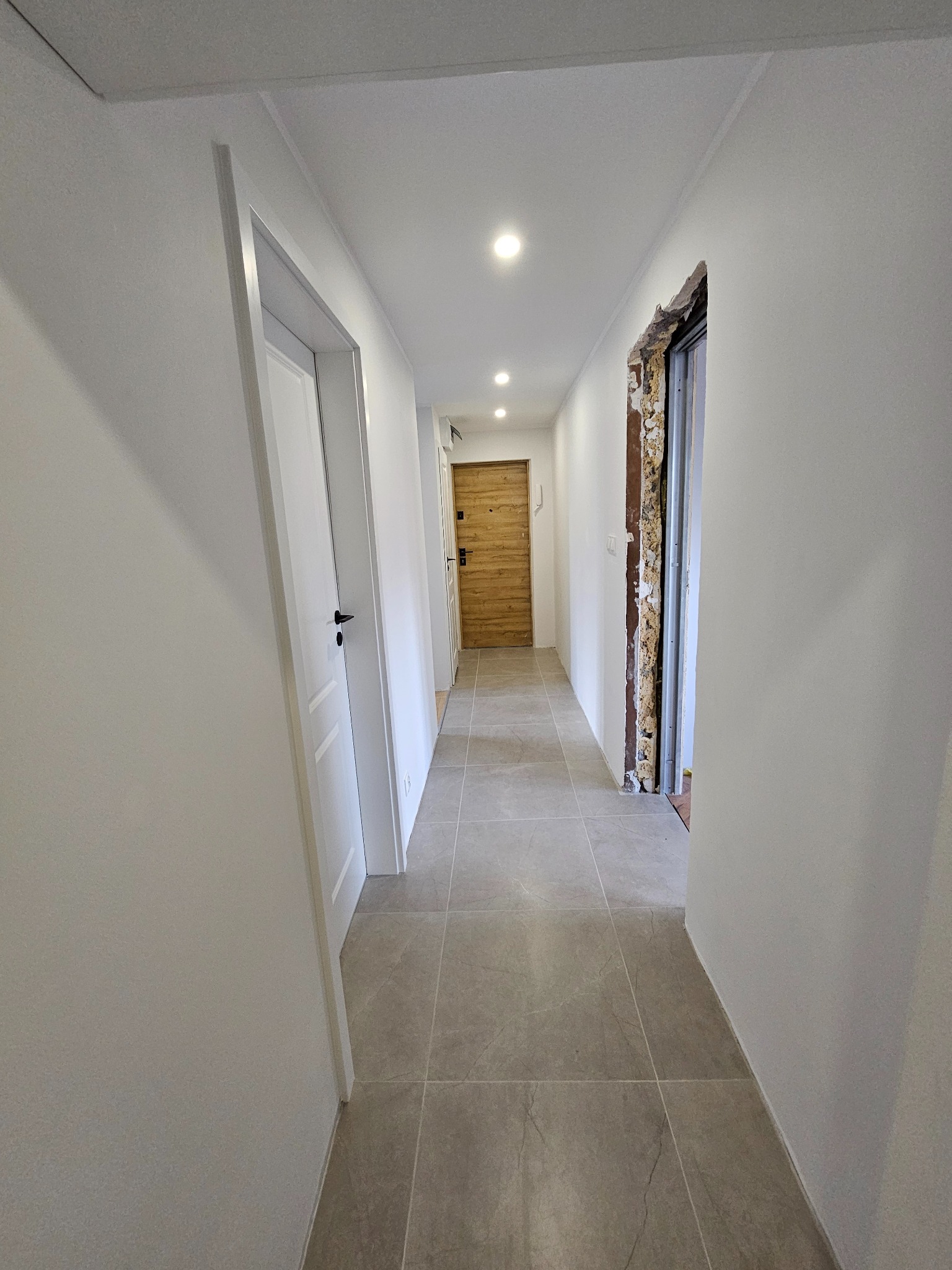Korytarz po remoncie: białe ściany, szara podłoga z płytek, białe drzwi po lewej, surowy otwór drzwiowy po prawej. W tle widoczna drewniana okładzina drzwi.