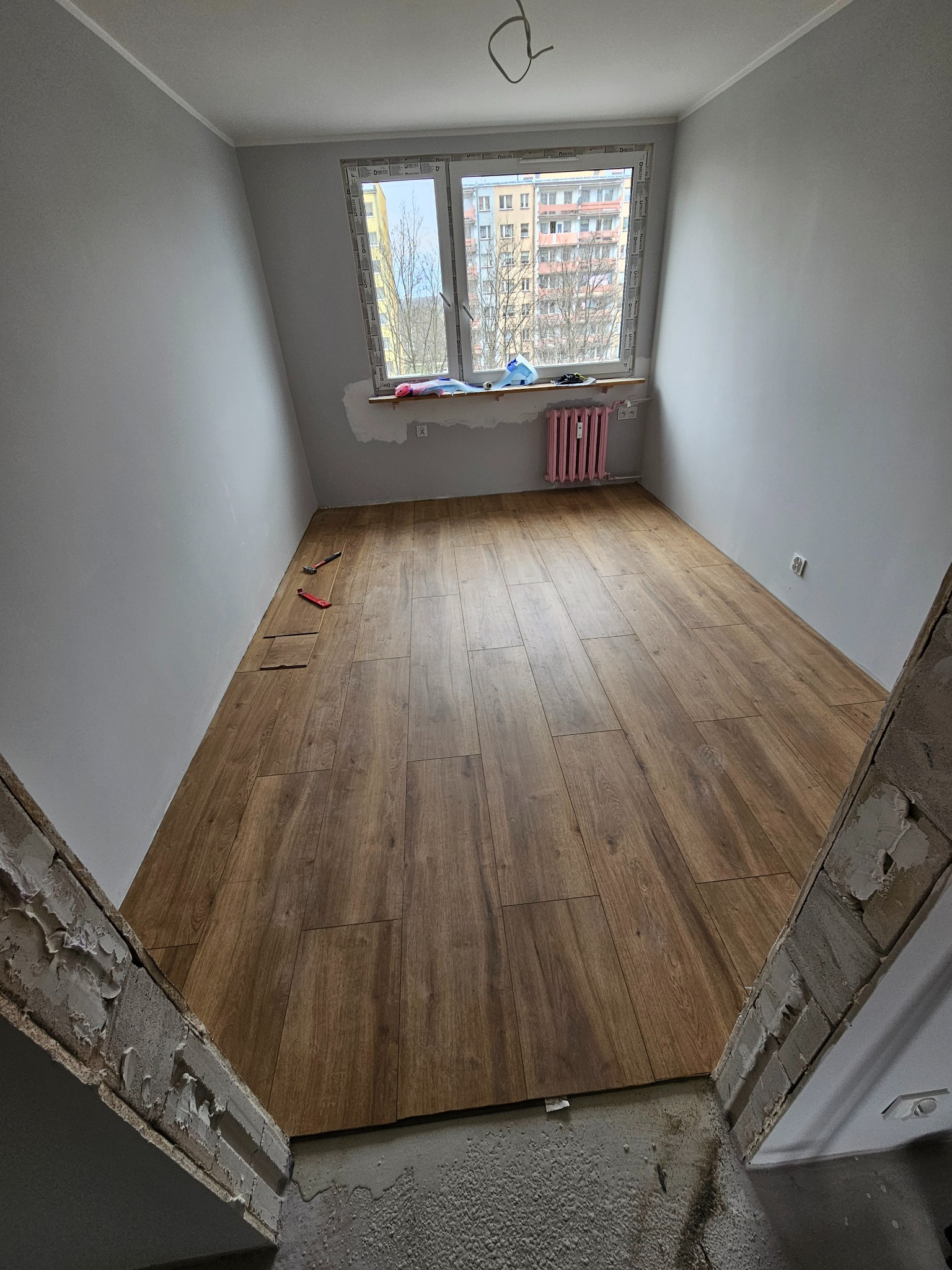 Ułożone panele podłogowe w pokoju z szarymi ścianami i różowym grzejnikiem pod oknem. Widoczne narzędzia do montażu paneli i niedokończone wykończenie przy progu.